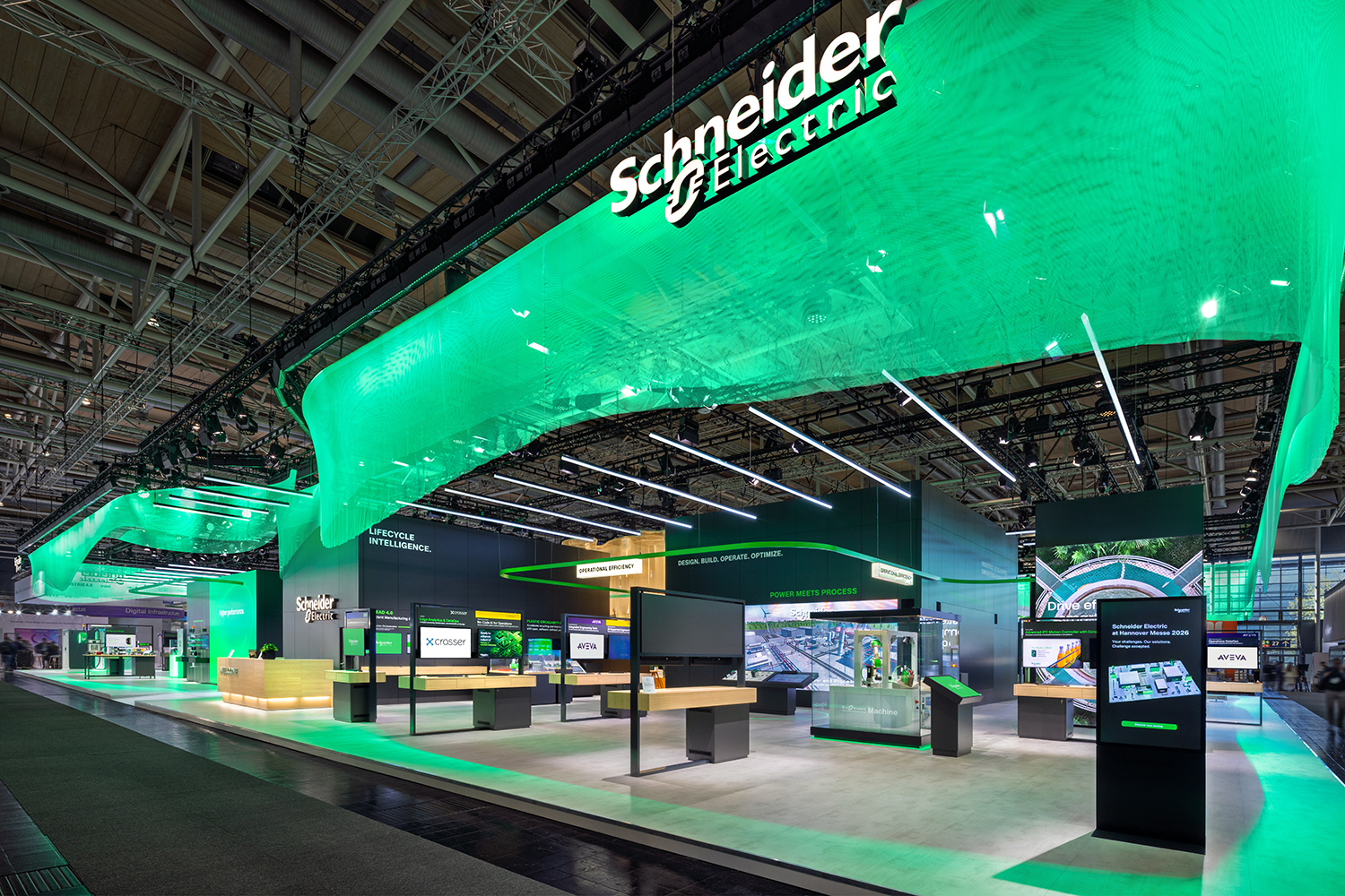 Weitwinkelansicht des doppelstöckigen Schneider Electric Messestands auf der HMI 2026 mit grüner Stoff-Lichtdecke und interaktiven Exponat-Tischen.