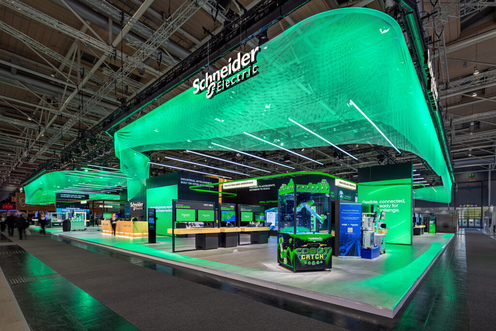 Corner view of the Schneider Electric HMI 2026 stand featuring the Cobot Catch robotic arm exhibit and green illuminated brand ceiling wave.