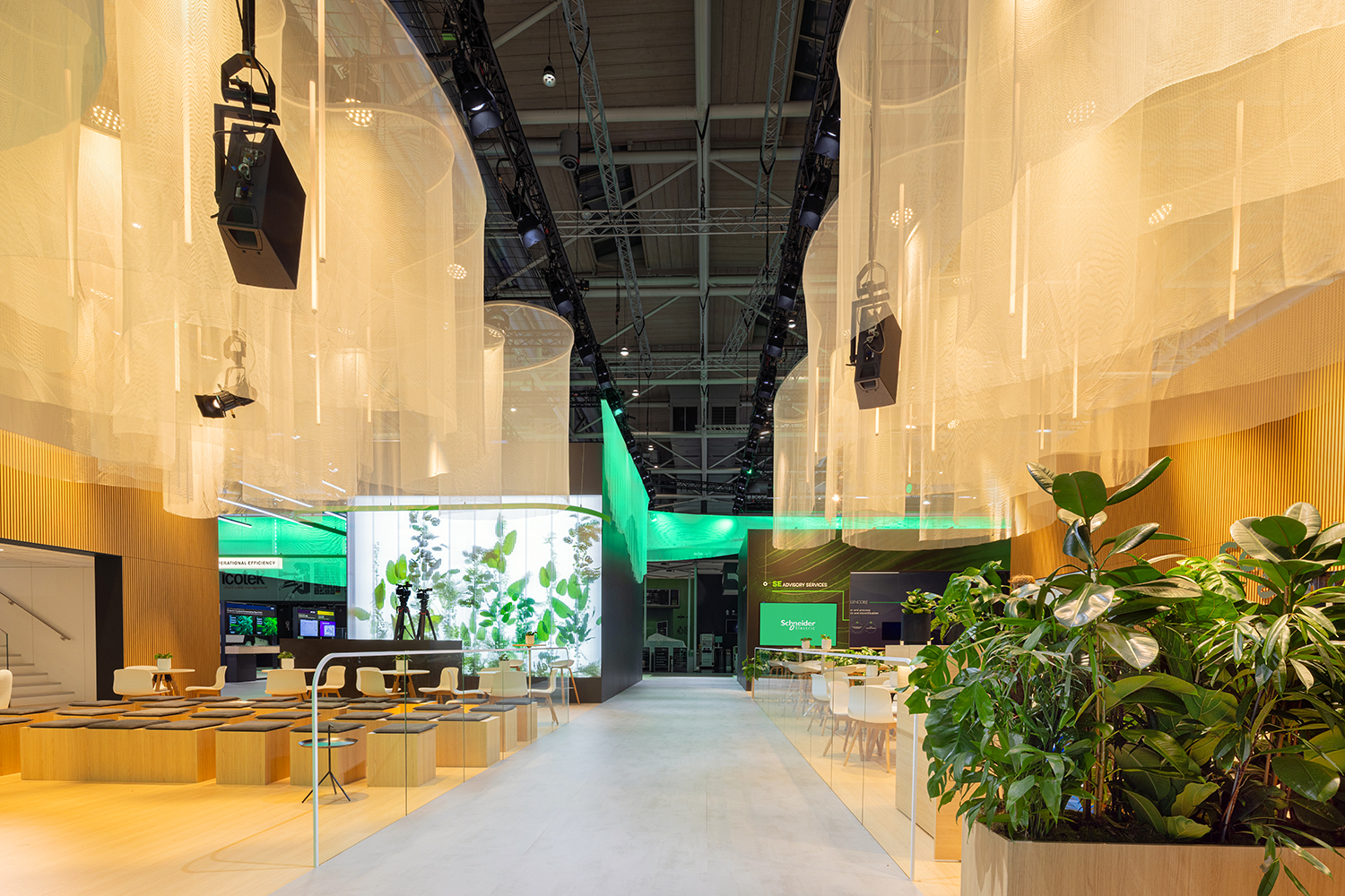 Lounge and presentation area featuring light wood flooring, mesh textile ceiling drapes, live plants, and modular seating.