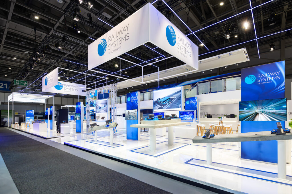 Open exhibition stand design for voestalpine Railway Systems featuring suspended ceiling structures and product display islands at InnoTrans 2026.